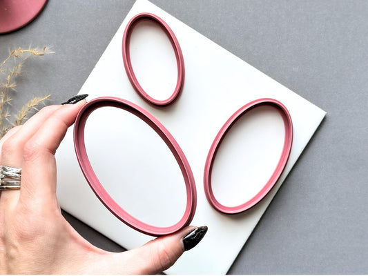 Three oval polymer clay cutters displayed in graduated sizes on a white tile, with the largest cutter held by hand to show the elongated oval shape and smooth cutting edge.