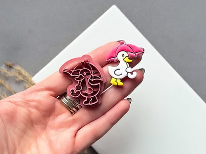 polymer clay cutter in the shape of a duck holding an umbrella, this cutter is for making polymer clay jewellery and accessories. The cutter is shown in a hand for scale along side a finished clay cut out earrinng.