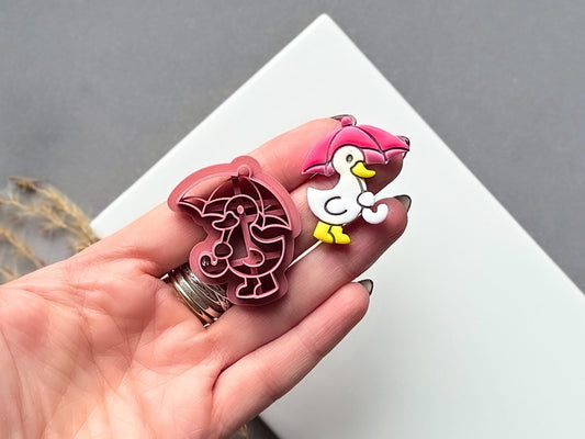 polymer clay cutter in the shape of a duck holding an umbrella, this cutter is for making polymer clay jewellery and accessories. The cutter is shown in a hand for scale along side a finished clay cut out earrinng.