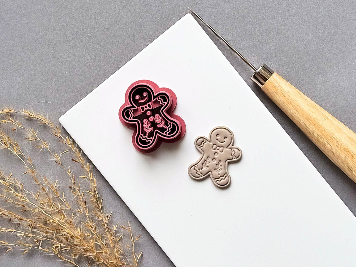 Gingerbread man polymer clay cutter displayed beside a matching finished clay embellishment, featuring a smiling face, bow tie, buttons, wavy icing details and decorative floral motifs on a white tile.