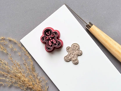 Gingerbread man polymer clay cutter displayed beside a matching finished clay embellishment, featuring a smiling face, bow tie, buttons, wavy icing details and decorative floral motifs on a white tile.