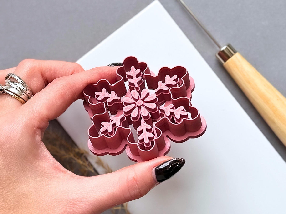 Large snowflake polymer clay ornament cutter held between fingers to showcase the raised stamp details, deep cutting walls and intricate six-branch design with a central flower and decorative leaf motifs.