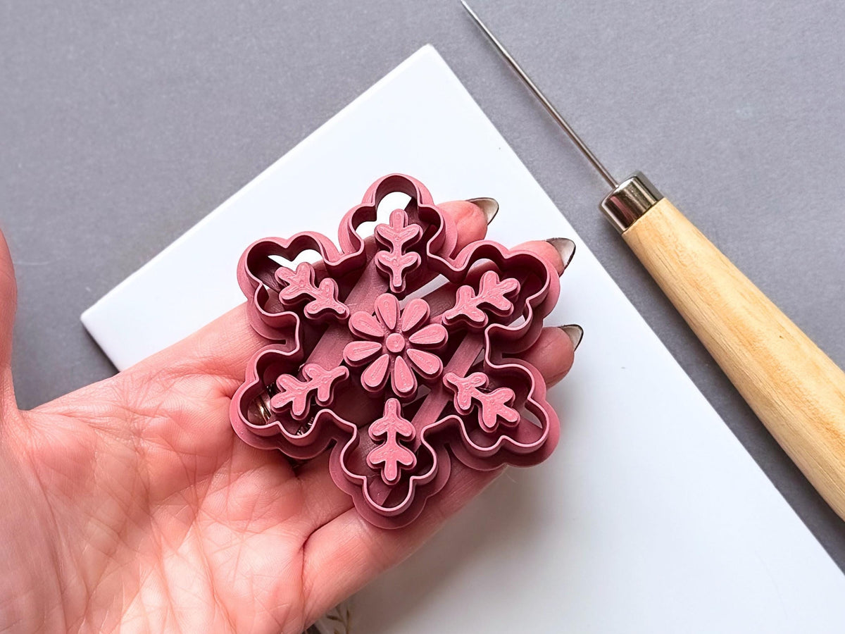 Large snowflake polymer clay ornament cutter held in a hand for scale, featuring a central flower and six decorative branches with leaf-like details, showcasing the raised stamp design and shaped cutting edge.