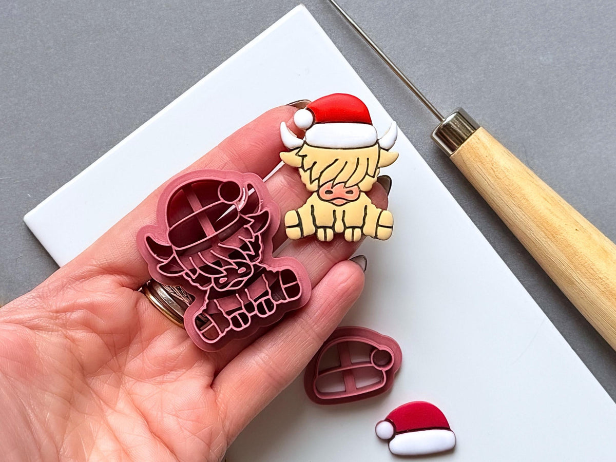 Highland cow Christmas polymer clay cutter and matching finished clay embellishment held in a hand for scale, with a separate Santa hat cutter and finished hat visible nearby.