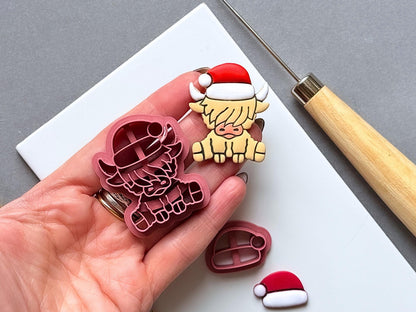 Highland cow Christmas polymer clay cutter and matching finished clay embellishment held in a hand for scale, with a separate Santa hat cutter and finished hat visible nearby.