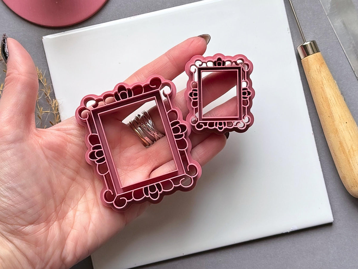 Two vintage-style rectangular frame polymer clay cutters in different sizes, held in a hand to show scale, featuring ornate decorative borders with loops, scallops and rounded accents.