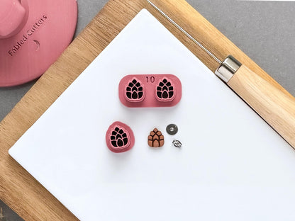 Pine cone polymer clay stamp cutter set displayed on a white tile, featuring two matching pine cone stamp designs in a joined holder and a separate single cutter, alongside a finished clay pine cone embellishment and small metal findings for scale.