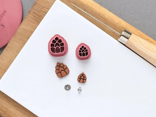 15 & 10 mm mini pink pinecone clay cutters with polymer clay cut out samples and earring posts on a white tile.