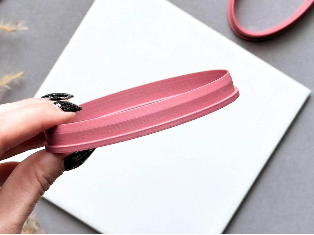 Oval polymer clay cutter held side-on to showcase its raised cutting wall, sturdy support base and elongated profile, with another oval cutter visible in the background.