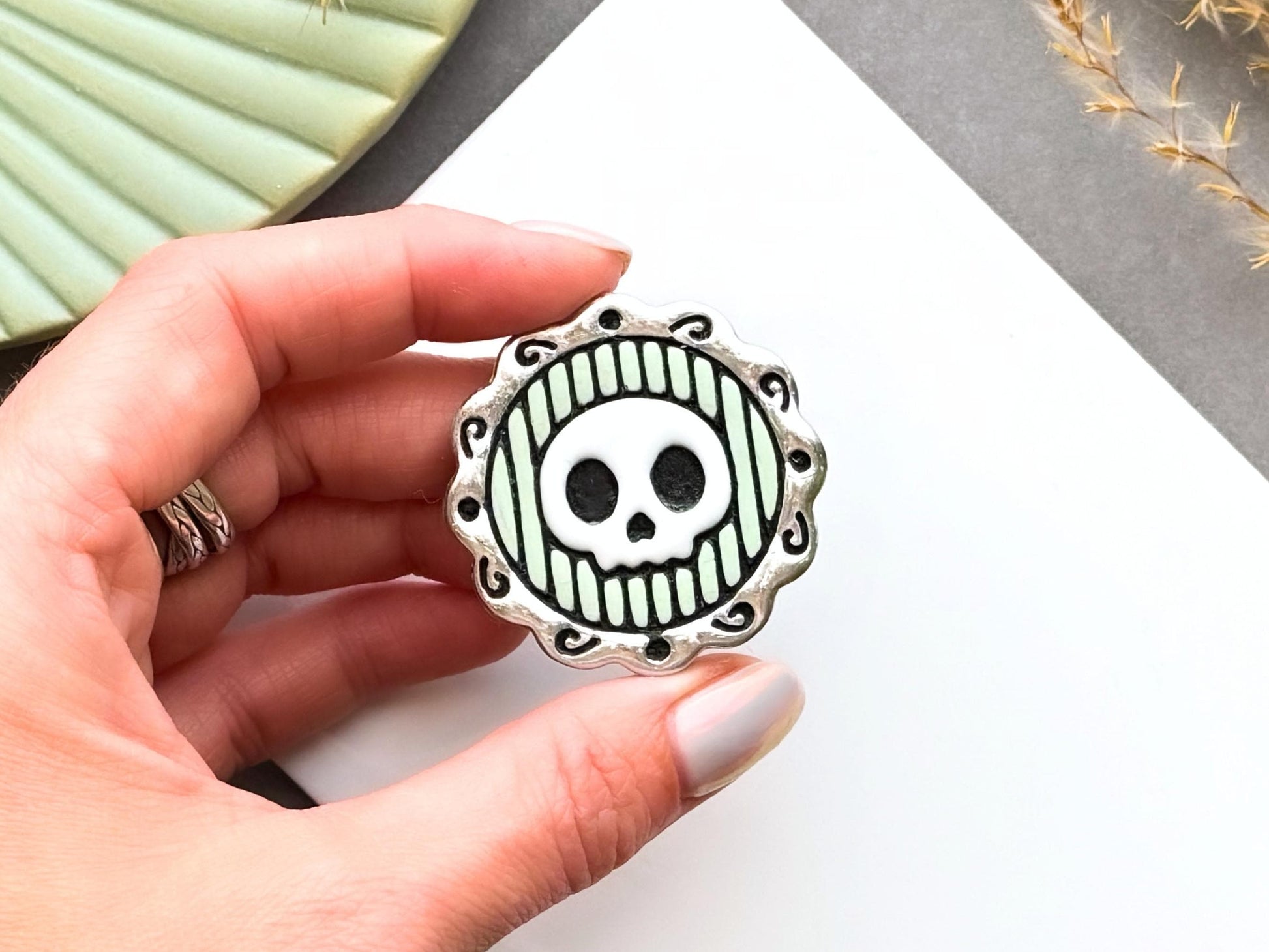 Finished polymer clay skull cameo shown being held in a hand to demonstrate scale, featuring a stylised skull within an ornate decorative frame for creating gothic jewellery, Halloween accessories and spooky craft projects.