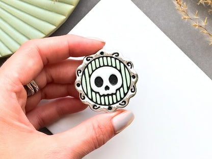 Finished polymer clay skull cameo shown being held in a hand to demonstrate scale, featuring a stylised skull within an ornate decorative frame for creating gothic jewellery, Halloween accessories and spooky craft projects.