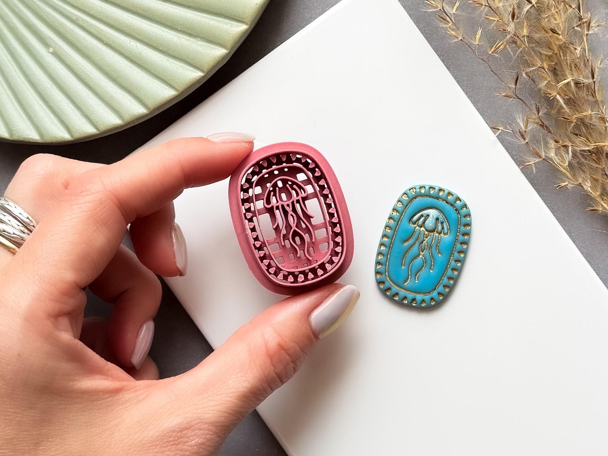 Jellyfish polymer clay cutter shown being held in a hand to demonstrate scale and intricate detail, displayed alongside a finished polymer clay jellyfish sample with a decorative beaded oval frame for creating ocean-inspired jewellery, charms and coastal craft projects.