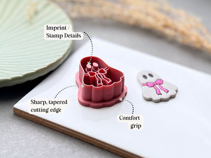 A ghost-shaped clay cutter in dusty pink with printed white and pink details, shown beside a matching white ghost polymer clay earring on a white plate. Text callouts point to the cutter's stamp details, sharp tapered cutting edge, and comfort grip.