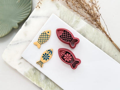 Two pink coloured clay cutters in the shape of fish with simple outline shape, one has a folk art style floral pattern with a big flower stamp in the middle, the other has scales detail. Next to them are polymer clay cut out samples in beige with stamped details painted blue.