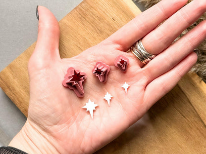 Three micro star polymer clay cutters in graduated sizes held in a hand for scale, alongside matching white clay cut-outs showcasing the distinctive four-point star shape with elongated lower points.
