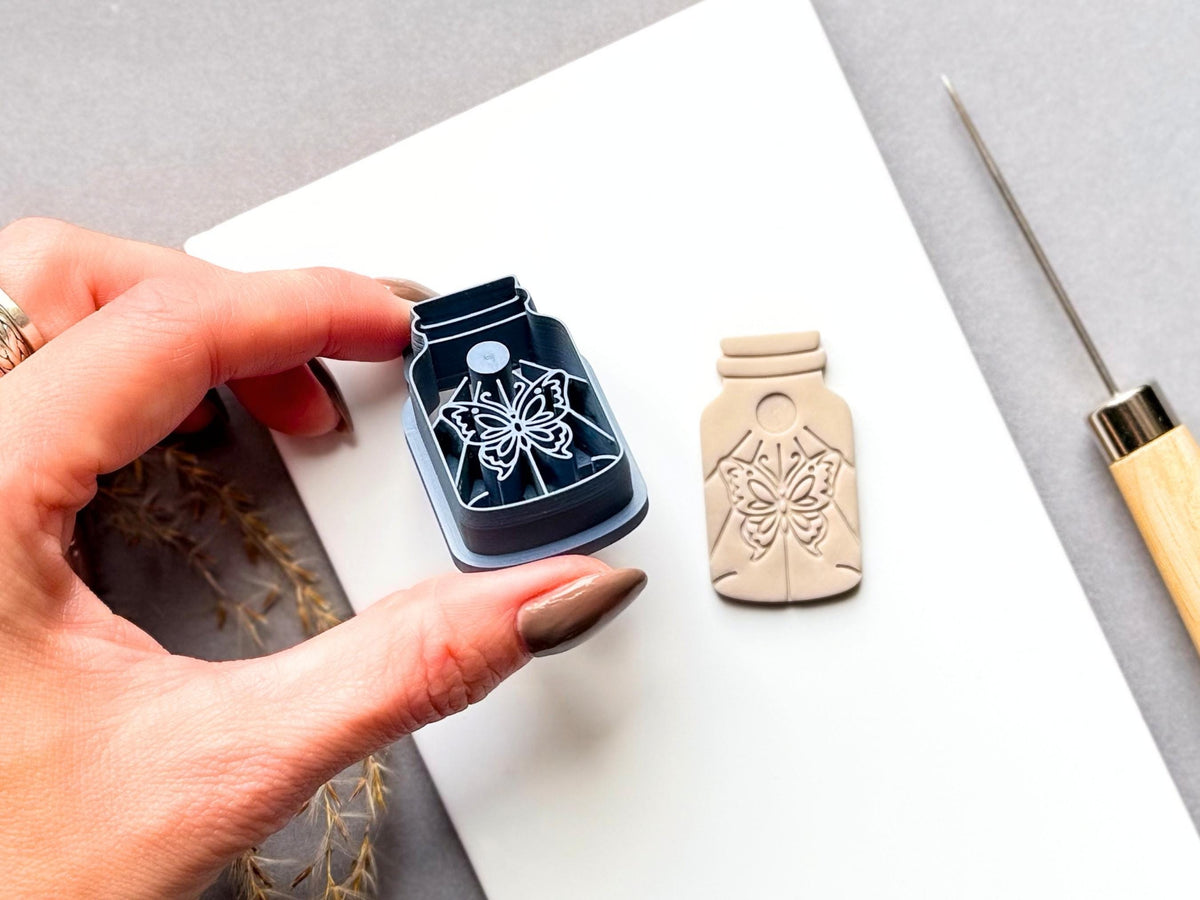 Butterfly specimen jar clay cutter held in a hand beside a finished cream polymer clay butterfly cut-out, featuring a detailed symmetrical butterfly design with decorative floral details. Blue 3D-printed cutter shown close-up to highlight the intricate stamp design.