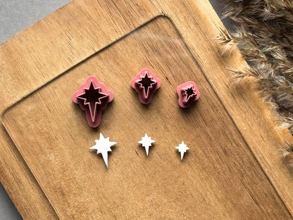 Three North star polymer clay cutters displayed in graduated sizes on a wooden surface, alongside matching white clay cut-outs showing the distinctive four-point star shape with elongated lower points.