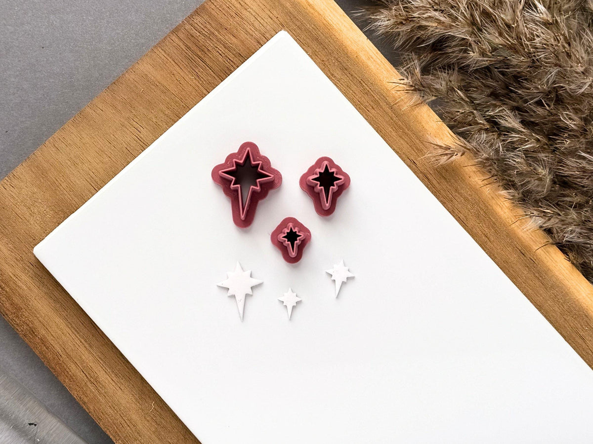 Three miniature star polymer clay cutters displayed in graduated sizes on a white tile, alongside matching white clay cut-outs featuring a four-point star design with elongated lower points.