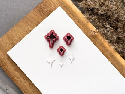 Three miniature star polymer clay cutters displayed in graduated sizes on a white tile, alongside matching white clay cut-outs featuring a four-point star design with elongated lower points.
