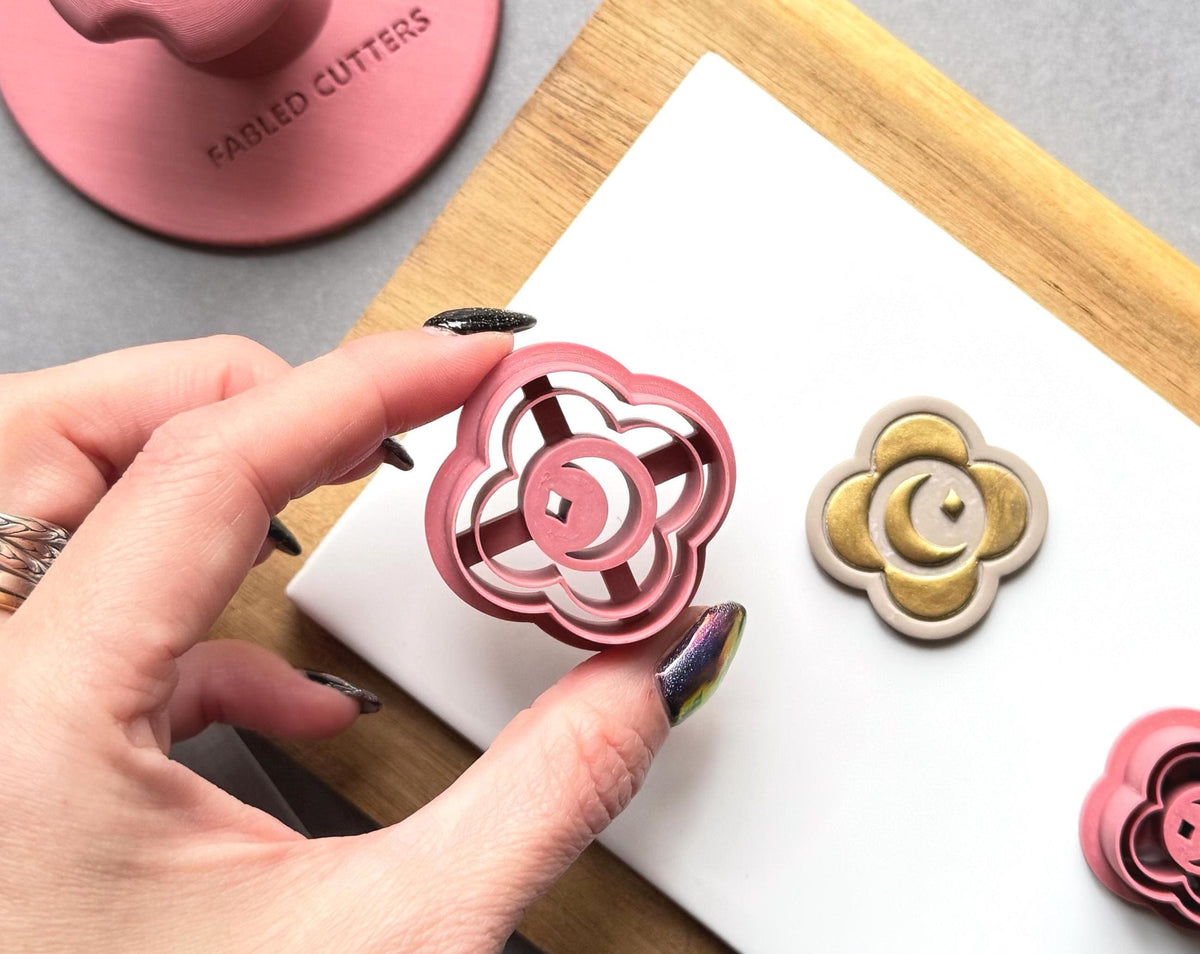 Floral crescent moon medallion polymer clay cutter displayed beside a matching gold-finished clay embellishment, with a smaller matching cutter in the background.