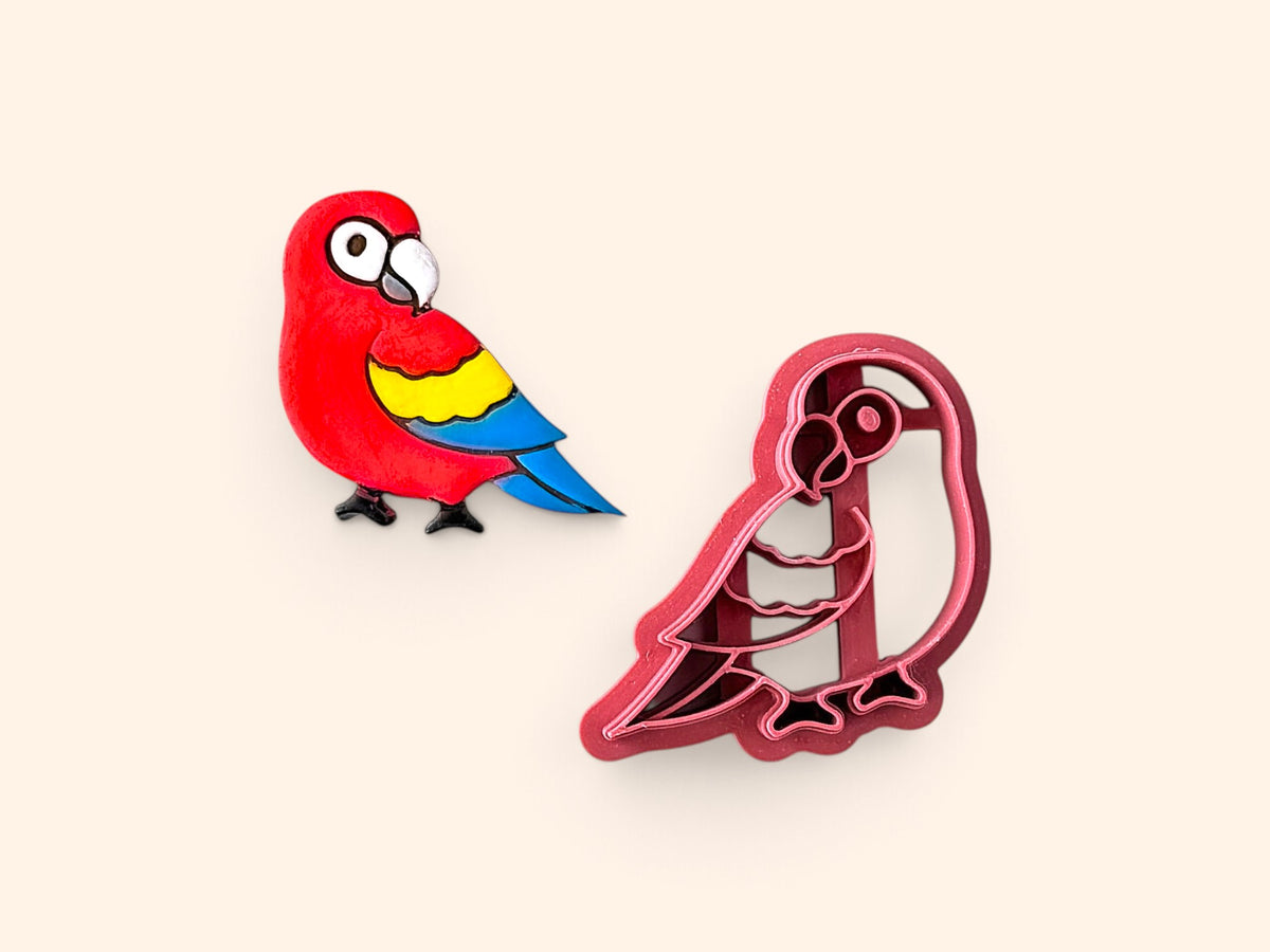 A small colorful parrot-shapedclay embellishment next to a matching pink plastic clay cutter, shown on a plain warm beige background.