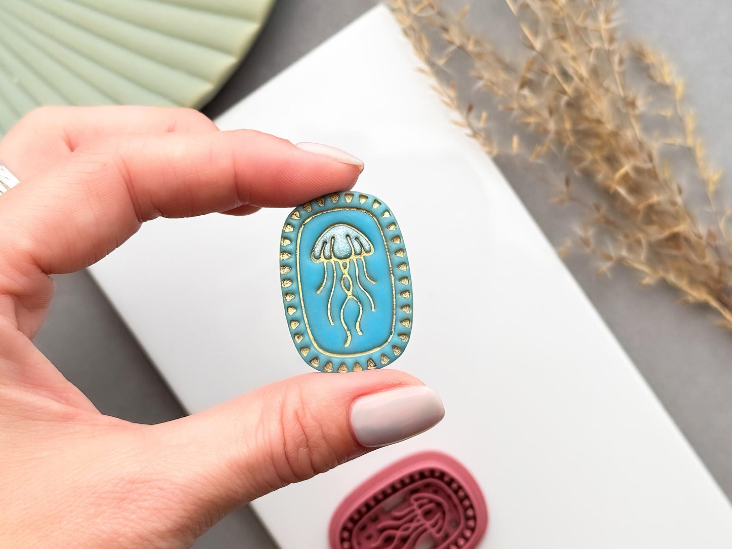 Finished polymer clay jellyfish charm shown being held in a hand to demonstrate scale, featuring a detailed jellyfish design within a decorative beaded oval frame for ocean-inspired jewellery and coastal craft projects.