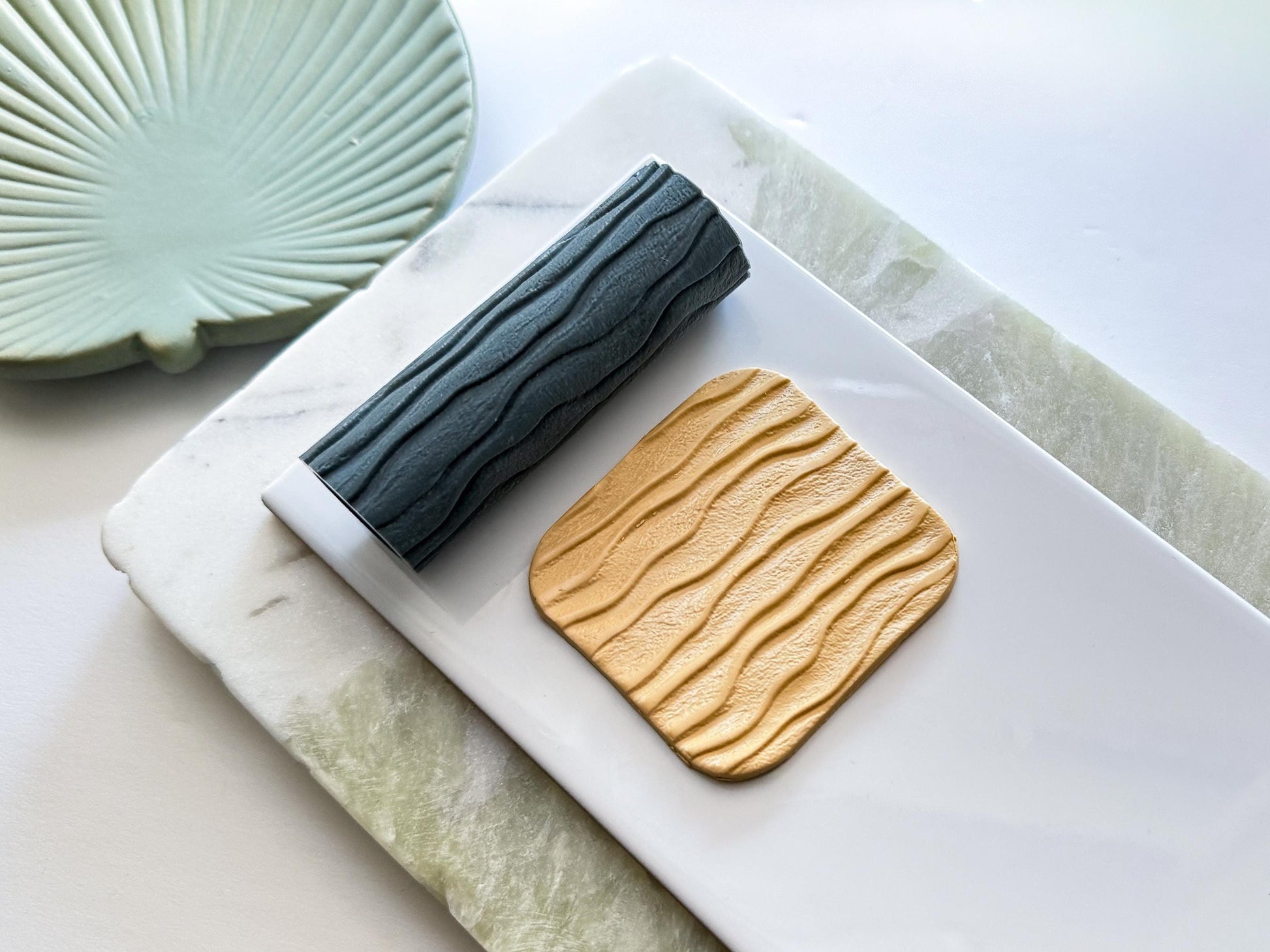 Sandy beach themed texture rolling pin with wavy horiztonal lines and sand texture. Next to the roller is a polymer clay cut out with the sandy beach texture on the surface, cut into a rounded square shape. Both are on a white and green tile set.