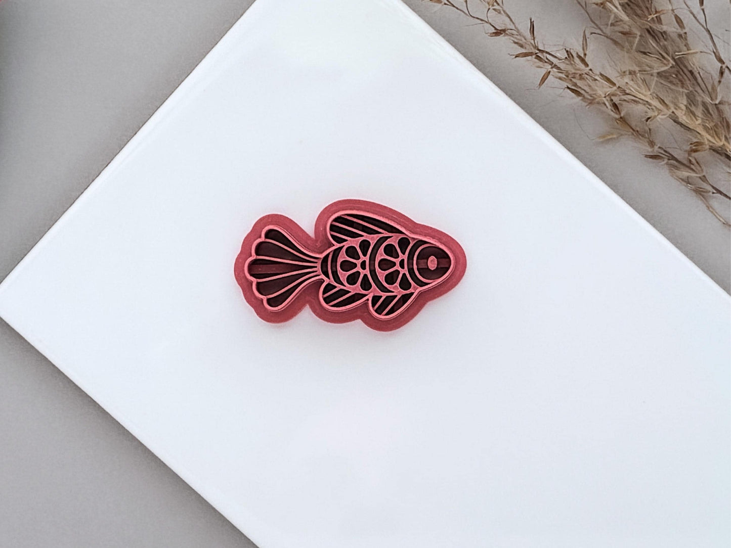 Ornamental fish polymer clay cutter with decorative scale and fin details, displayed on a white ceramic tile. Folk art-inspired fish design for jewellery making and craft projects