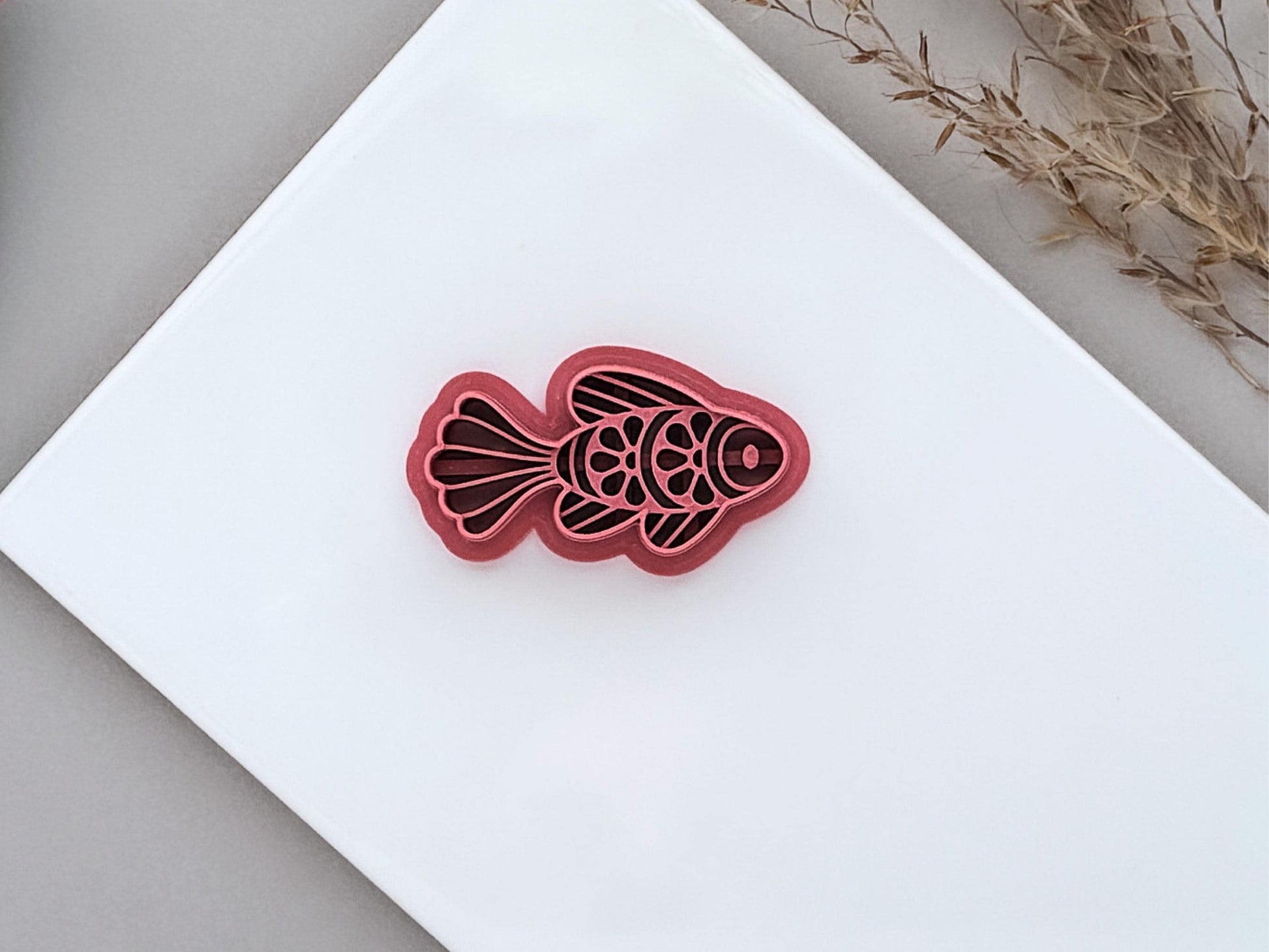 Ornamental fish polymer clay cutter with decorative scale and fin details, displayed on a white ceramic tile. Folk art-inspired fish design for jewellery making and craft projects