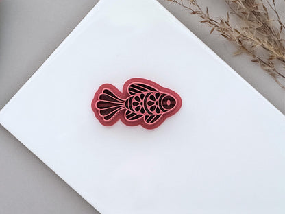 Ornamental fish polymer clay cutter with decorative scale and fin details, displayed on a white ceramic tile. Folk art-inspired fish design for jewellery making and craft projects