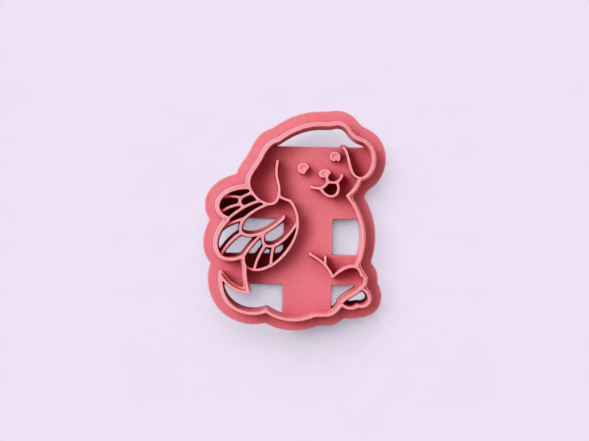 A pink plastic polymer clay cutter shaped like a cartoon dog with fairy wings, shown on a solid light lavender background.