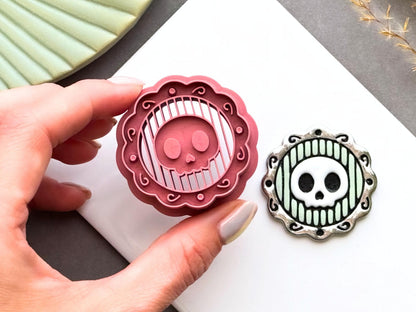 Skull cameo polymer clay cutter shown being held in a hand to demonstrate scale and intricate detail, displayed alongside a finished polymer clay skull cameo sample with an ornate decorative frame for creating gothic jewellery, Halloween charms and spooky craft projects.