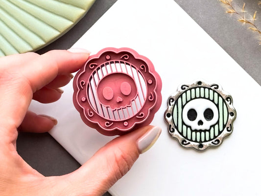 Skull cameo polymer clay cutter shown being held in a hand to demonstrate scale and intricate detail, displayed alongside a finished polymer clay skull cameo sample with an ornate decorative frame for creating gothic jewellery, Halloween charms and spooky craft projects.