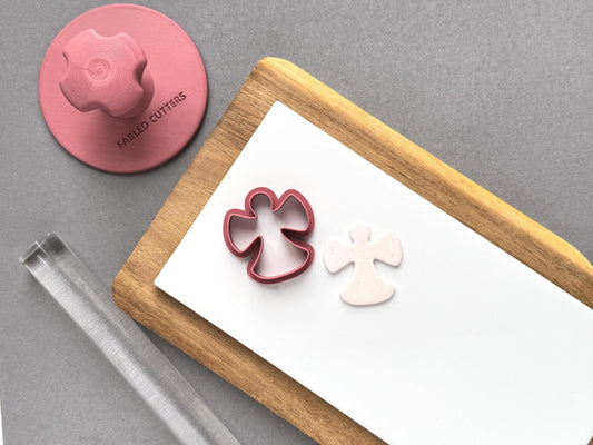 Snow angel polymer clay cutter displayed beside a matching sample clay cut-out on a white tile, with polymer clay tools arranged around the workspace.